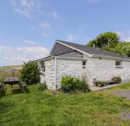 A white stone cottage with a dark roof and a wooden picnic table outside on the grass at Y Bwthyn in Elidir Fach Mountain near Llanberis