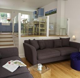 A living room with two brown sofas a magazine on one and a coffee cup on a glass table with a blue bean bag and a dining area with chairs and blue cabinets at 3 Eddystone Row in Salcombe