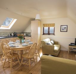 A kitchen dining area with a round wooden table set for six people and two sofas by a window at 4 Armada House in Hope Cove