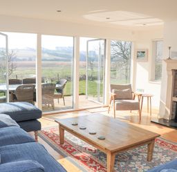 A living room with a wooden coffee table blue sofas a rug and a beige armchair next to a fireplace with glass doors opening to a garden at Sea Peep in Thurlestone