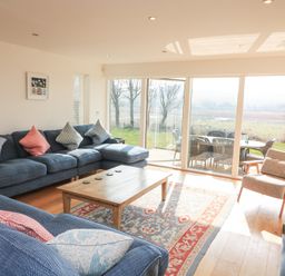 A living room with blue sofas cushions a wooden coffee table and armchair next to large glass doors at Sea Peep in Thurlestone