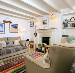 A living room with a sofa, armchair, colorful striped rug, fireplace, bookshelf, and framed pictures at Bow Cottage in East Portlemouth