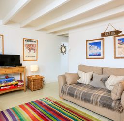 A living room with a beige sofa with cushions a colorful striped rug a wooden TV stand with games and a lamp at Bow Cottage in East Portlemouth