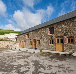 An exterior view of a stone building with multiple wooden doors and windows and flower pots outside at Butterwell Barn Dartmouth