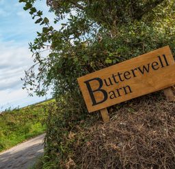 A wooden sign with Butterwell Barn written on it near a country road surrounded by bushes at Butterwell Barn in Dartmouth