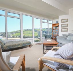 A sitting room with chairs a sofa a coffee table and a bookcase with large windows overlooking green hills at Edinburgh House in Salcombe
