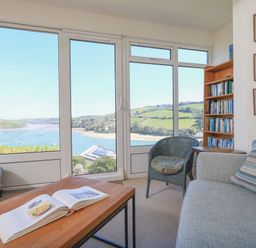 A living room with a bookshelf sofa chair and coffee table with an open book by large windows overlooking a river and hills at Edinburgh House in Salcombe