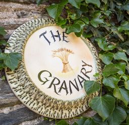 A circular ceramic sign with the words The Granary and a bundle of wheat surrounded by green ivy on a stone wall at The Granary Malborough