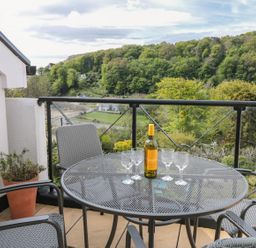 A balcony with a round metal table and four chairs with a bottle and four glasses on the table overlooking a wooded hillside at The Hoot 16 St Elmo Court in Salcombe