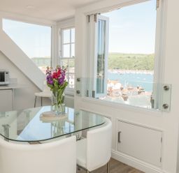 A dining area with a glass table and white chairs next to a large window overlooking a harbor at Kings View in Dartmouth
