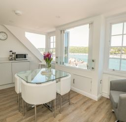 A kitchen dining area with glass table white chairs grey armchair and windows overlooking water at Kings View in Dartmouth
