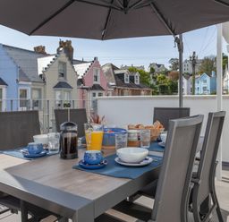 An outdoor dining table with chairs set for breakfast under a large umbrella on a balcony with colorful houses in the background at Lower Fernlea in Salcombe