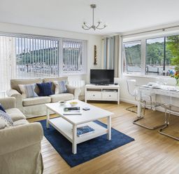 A living room with two beige sofas a white coffee table a TV on a stand and a dining table with transparent chairs by large windows at Leeside Dartmouth
