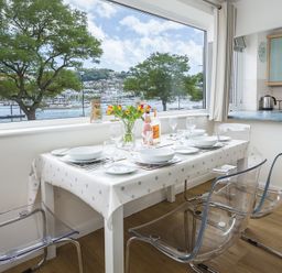 A dining area with a table set for four and clear chairs by a large window overlooking a waterfront at Leeside in Dartmouth
