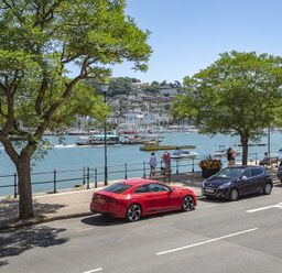 A waterfront street with parked cars and trees overlooking a harbor with boats and hills in the background at Leeside in Dartmouth