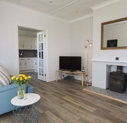 A living room with a blue armchair, small white table with yellow flowers, television on a wooden stand, wood stove in a tiled fireplace, and an open door to a kitchen at Seaspray Bigbury-on-Sea