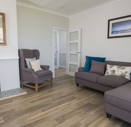 A living room with a gray armchair and sectional sofa with cushions next to a fireplace and a wall mirror at Seaspray Bigbury-on-Sea