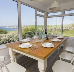 A dining area with a wooden table set with plates and glasses surrounded by white chairs with large windows showing a coastal view at Seaspray Bigbury-on-Sea