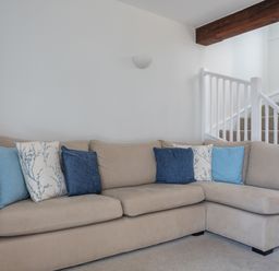 A beige L-shaped sofa with blue and patterned cushions in a living room near a staircase with white railing and a wooden beam at Stable End Cottage in Malborough