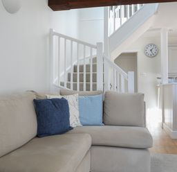 A living room with a beige sectional sofa with blue and white cushions near a white staircase and kitchen at Stable End Cottage in Malborough