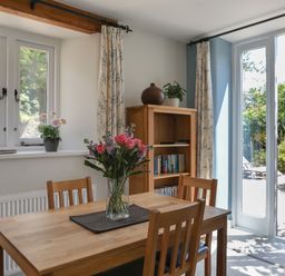 A dining area with a wooden table and four chairs a vase with flowers on the table a bookcase with books and plants curtains on windows and glass doors leading outside at Stable End Cottage Malborough