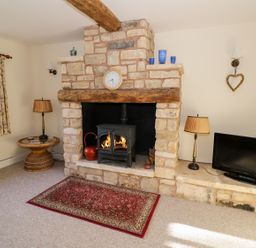 A living room with a stone fireplace with a wood stove and clock above it a small table with a lamp and floral curtains and a television on the hearth at The Hideaway in Eckington