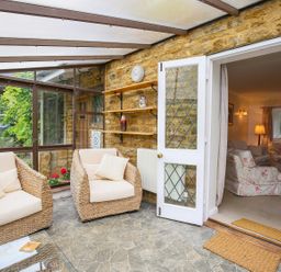 A sunroom with wicker armchairs and cushions next to a doorway leading to a living room at Stone Wheel Cottage in Hook Norton
