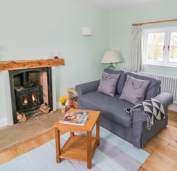 A living room with a fireplace, a gray sofa with cushions, a wooden coffee table with magazines, and a window at Wigrams Canalside Cottage in Napton-on-the-Hill