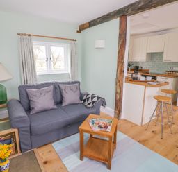 A living room with a blue sofa, wooden side table, green lamp, small wooden coffee table, and an open kitchen with two stools at Wigrams Canalside Cottage in Napton-on-the-Hill