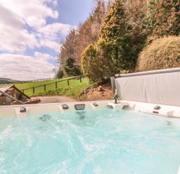 An outdoor hot tub with bubbling water near a hillside with grass, trees, a wooden fence, and a small stone building at Treboeth in Knighton