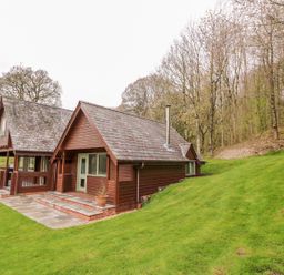 Two wooden cabins with slate roofs near a grassy hill and leafless trees at Pines Pitch in Knighton