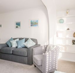 A living room with a gray sofa and armchair with cushions and white shelves with decorations at Bayview in Mevagissey
