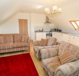 A living room and kitchen with a striped sofa, patterned armchairs, a wooden door, white cabinets, and skylight windows at Llain Haf in Llangefni