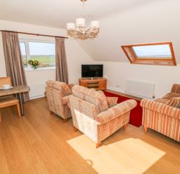 A living room with a dining table with chairs two patterned armchairs a striped sofa a television on a wooden stand and windows with curtains at Llain Haf in Llangefni