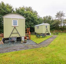 Two green shepherd huts with wooden stairs and benches on a grassy area with trees in the background at Shepherds Hut - The Hurdle in Rosemarket