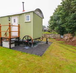 A green shepherds hut with wheels on a gravel area beside grass and trees at Shepherds Hut - The Hurdle in Rosemarket