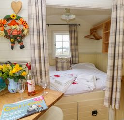 A small bedroom with a bed towels and curtains with a table holding flowers wine glasses and a magazine at Shepherds Hut - The Hurdle in Rosemarket
