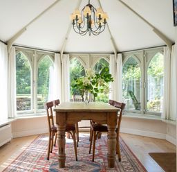 A dining area with a wooden table and four chairs under a chandelier in a room with arched windows and white curtains at All Souls Cottage in Eastleach Cotswolds