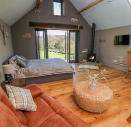 A room with a bed a wood burning stove a leather sofa and glass doors opening to a garden at Thompson Rigg Barn Langdale End near Thornton Dale