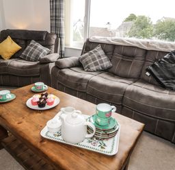 A living room with a wooden coffee table holding a tea set and cupcakes and two brown sofas with cushions at Ty Gilbert in Benllech