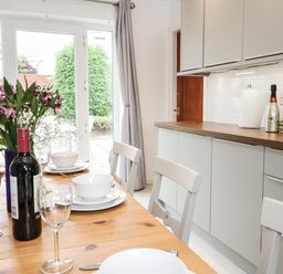 A dining area with a wooden table set with white plates bowls wine glasses and a bottle of wine next to a kitchen with grey cabinets and a countertop at Ty Gilbert in Benllech