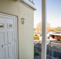 A white front door with a wall-mounted light next to a balcony with glass and metal railings overlooking a street and hills at 1 Lyndhurst in Salcombe