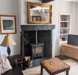 A living room with a wood-burning stove in a stone fireplace a wooden coffee table a TV on a wooden cabinet a lamp on a side table and a sofa with cushions at Rose Cottage in Lowick Bridge