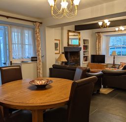 A dining area with a round wooden table and chairs next to a living room with sofas a fireplace and a television at Rose Cottage in Lowick Bridge