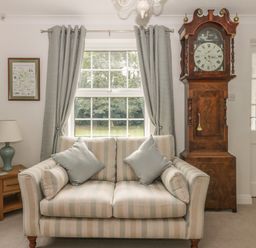 A sitting room with a striped sofa cushions a grandfather clock lamp on a wooden side table and curtains at Humble Bee Cottage in Wold Newton near Hunmanby