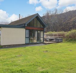 A house with large windows and a wooden deck with outdoor furniture on a grassy lawn near rocky hills at Restharrow Lodge in Bamburgh