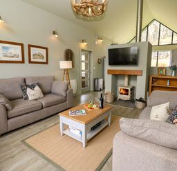 A living room with two sofas a coffee table a wood stove with a mounted TV and large windows at Restharrow Lodge in Bamburgh