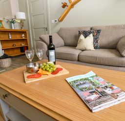A living room with a beige sofa two decorative cushions a wooden coffee table with a magazine a bottle of wine two wine glasses grapes and cheese at Restharrow Lodge Bamburgh