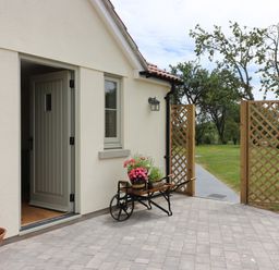 An outdoor patio area with a potted plant and a wheelbarrow holding flowers next to an open door and a wooden lattice gate at The Retreat in Lympsham