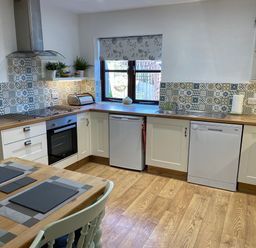 A kitchen with wooden flooring wooden countertops white cabinets a dining table with placemats and chairs at Stable Cottage in Shipham